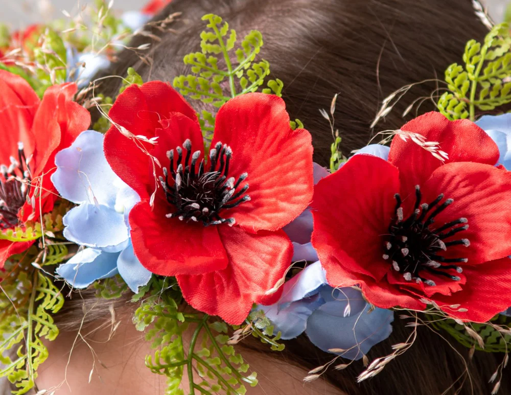 Bilde tuvplānā: mākslīgs vainags ar arkaniem, debessziliem ziediem un zaļumiņiem